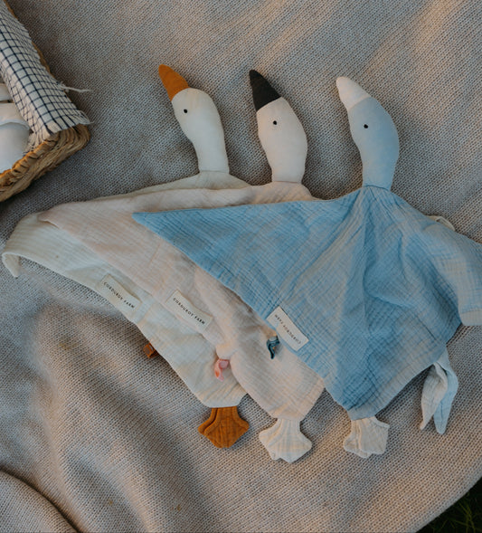 A soft fabric duck-shaped baby lovey or comforter laid on a beige blanket, with three plush duck heads and a light blue and white body. A small basket is partially visible at the upper left edge.