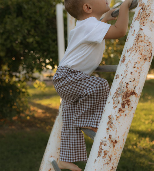 A toddler in a white t-shirt and brown-and-white gingham pants climbs a weathered white metal ladder outdoors on a grassy lawn.