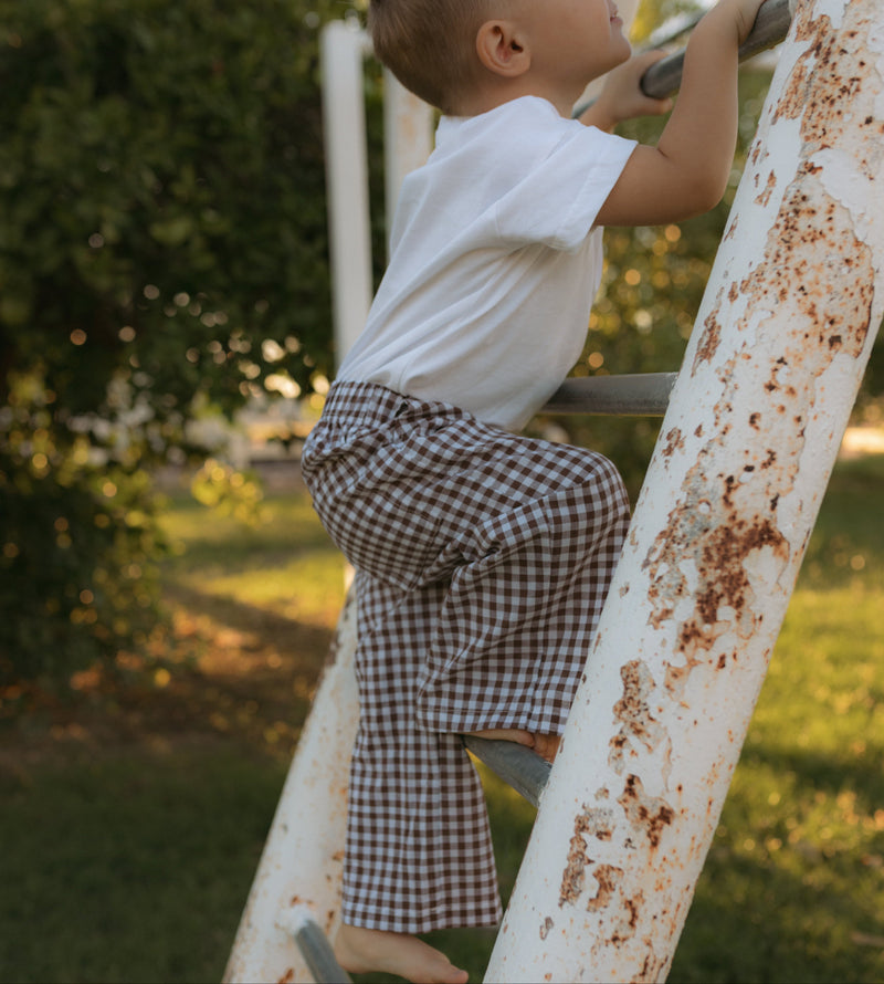 A toddler in a white t-shirt and brown-and-white gingham pants climbs a weathered white metal ladder outdoors on a grassy lawn.
